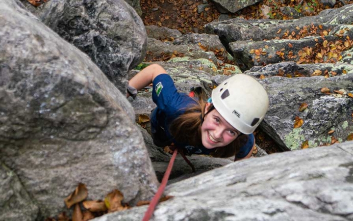 a girl looks up at the camera and smiles while rock climbing on an outward bound course